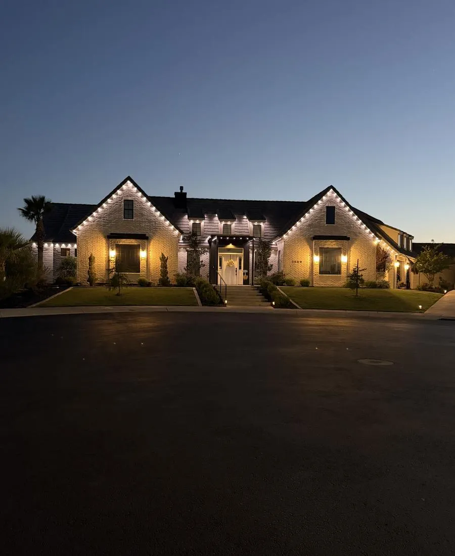 White brick two-story home at dusk with warm white permanent LED lighting outlining the gables, dormers, and entry
