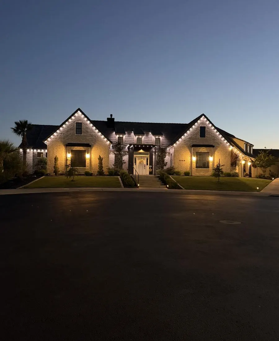 Two-story white brick home at dusk with warm white permanent LED lighting tracing both gables, the dormers, and the entry portico