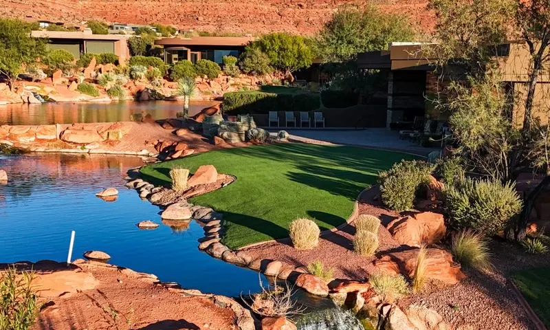 Artificial turf lawn and putting area beside a pond with red rock formations and a desert modern home in the background
