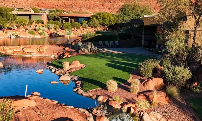 Artificial turf lawn and putting area beside a pond surrounded by red rock formations with a desert modern home in the background